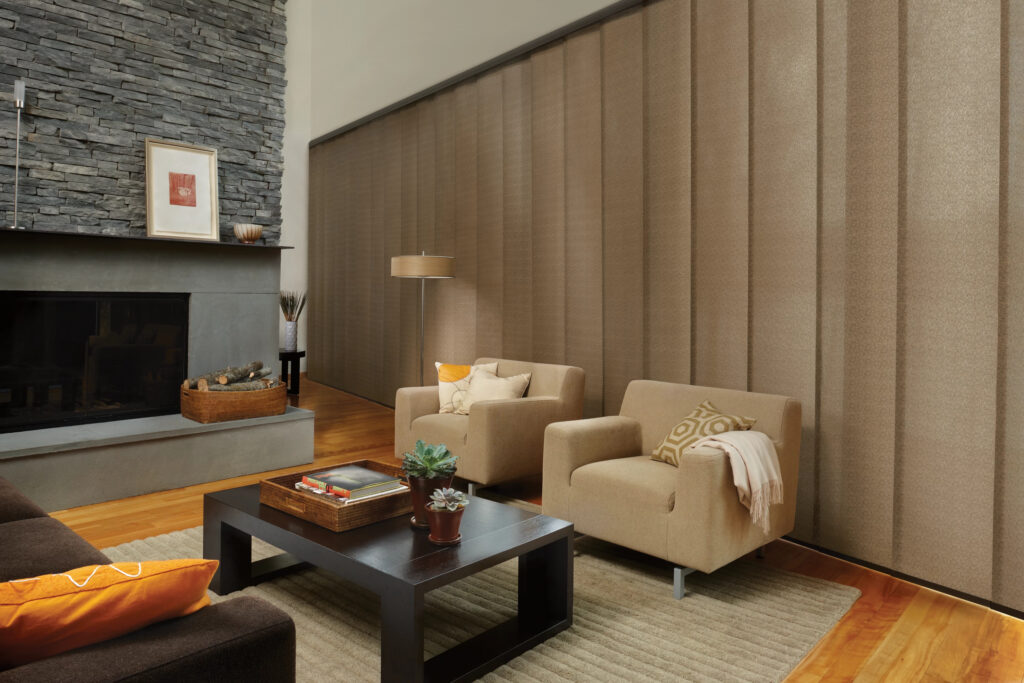 Modern living room with beige armchairs, dark wood coffee table, rug, stone fireplace, and decorative accents. One wall is covered by Skyline® Gliding Window Panels in beige, adding a sleek touch. Plants decorate the table and mantel.