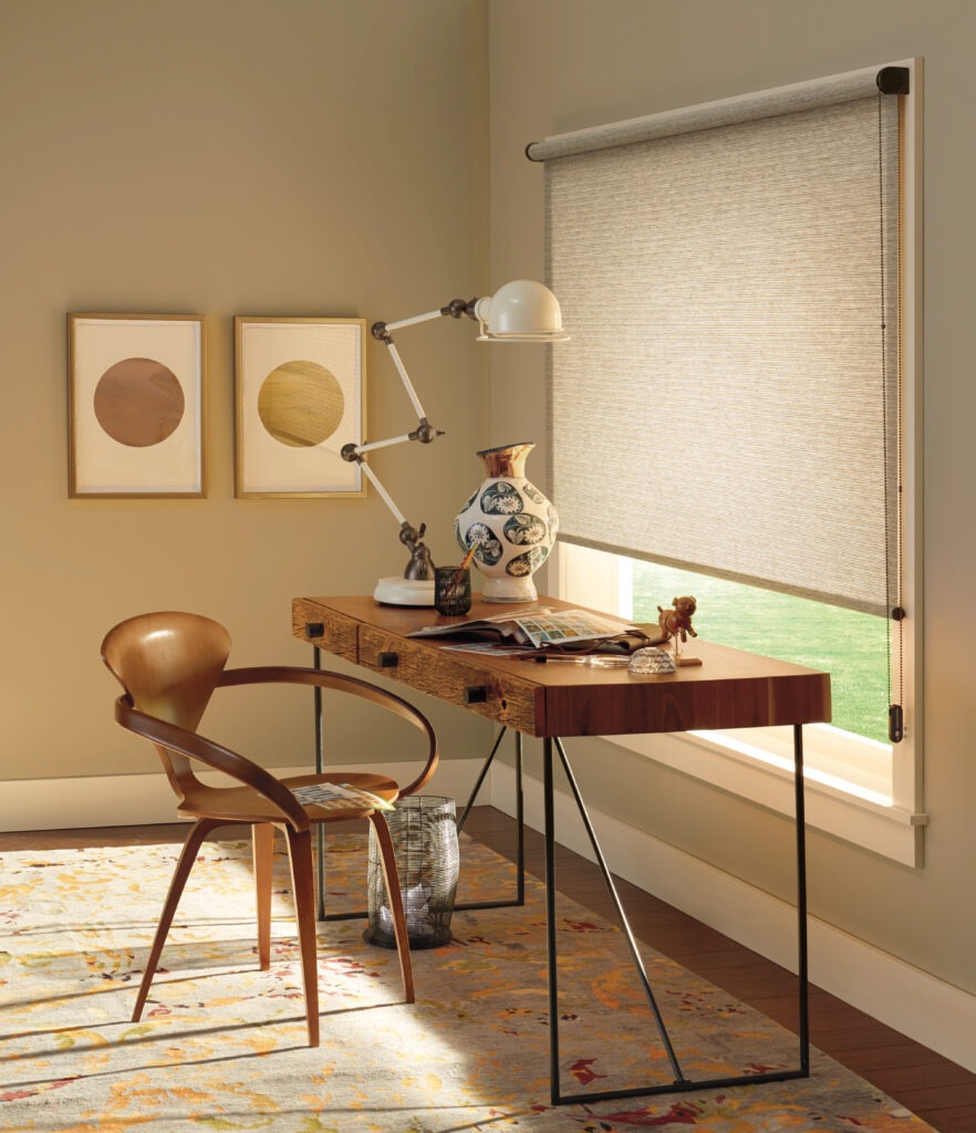A modern home office with a wooden desk, mid-century chair, adjustable lamp, decorative vase, framed art, and a large window featuring Designer Roller Shades in Paramus NJ that fill the space with natural light.
