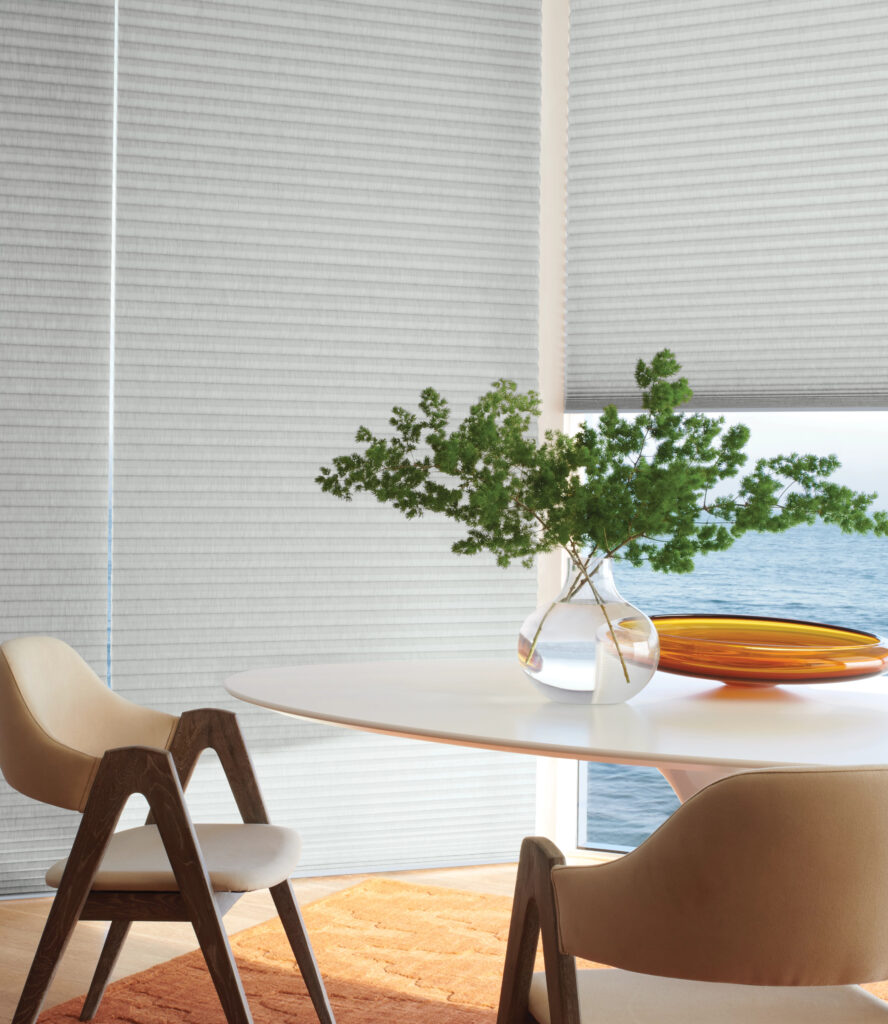 A round white table with a glass vase of green foliage and orange plates sits between two modern beige chairs on an orange rug, beside large windows featuring stylish window treatments in Morristown NJ overlooking water.