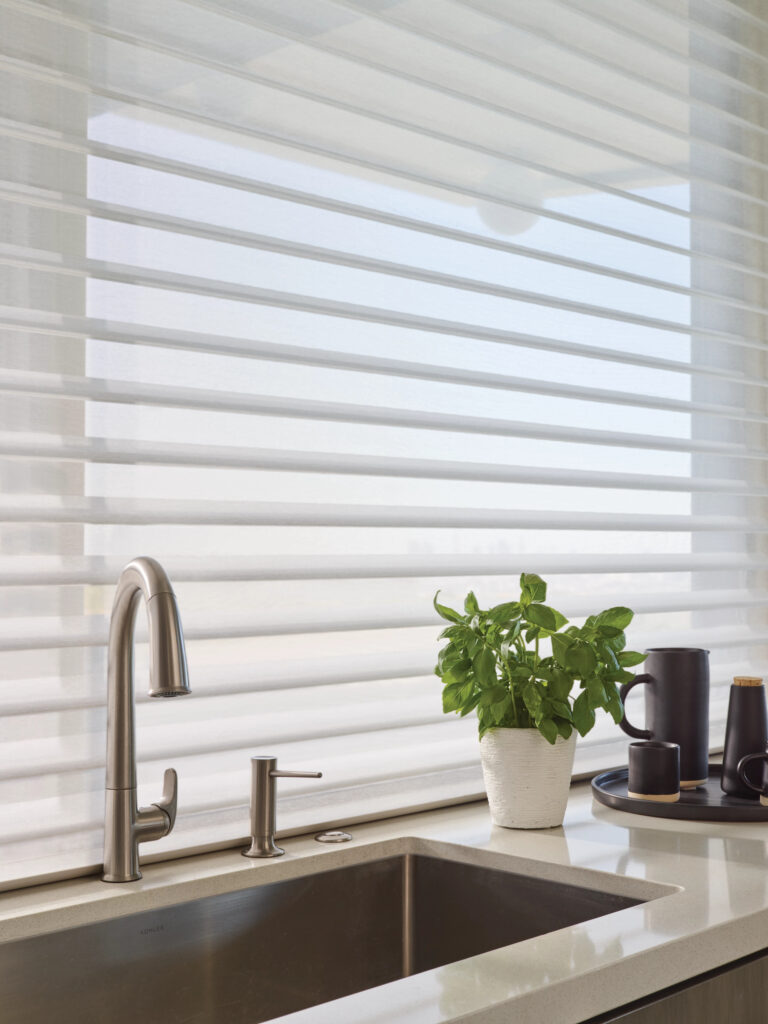 A modern kitchen sink with a stainless steel faucet and soap dispenser, a potted plant, and a tray with mugs sits in front of a large window adorned with elegant window blinds in Paramus NJ, letting in soft natural light.