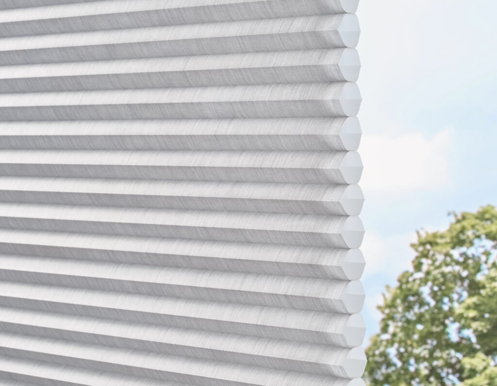 Close-up of a partially open Applause® Honeycomb Shade in white, softly filtering daylight. Perfect stylish window treatment for Paramus, NJ homes, with blurred green branches and blue sky visible outside.