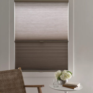 A window with partially open Duette Honeycomb Shades—ideal for stylish windows in Paramus, NJ—a woven chair sits in front, next to a small white table holding a flower arrangement, book, and glass of water.