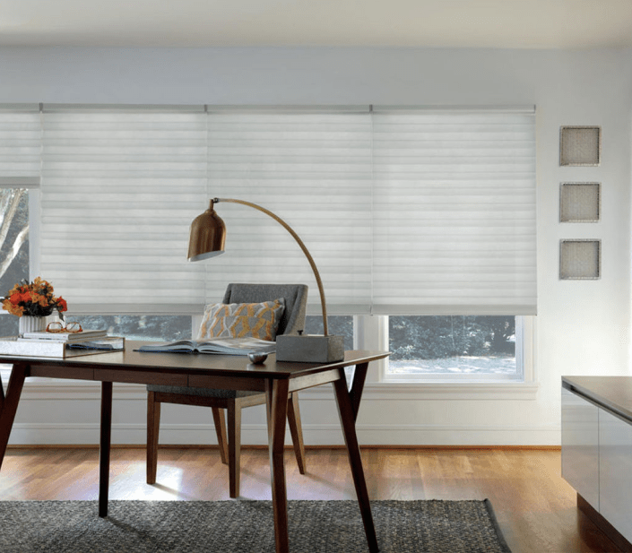 A modern home office with a wooden desk, chair, and gold lamp sits in front of large windows adorned with stylish blinds in Paramus NJ. A vase with flowers and books rests on the desk as light streams in, brightening the room.