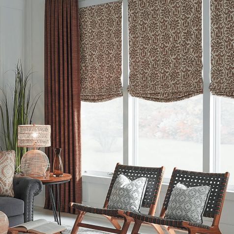 A bright living room in Paramus NJ features Custom Window Draperies on large windows, two wooden lounge chairs with decorative pillows, a side table with a lamp, and a corner plant.