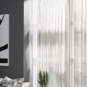 A modern living room corner in Paramus, NJ with Custom Window Draperies as sheer white curtains, a black-and-white abstract painting on a gray wall, a patterned cushion, and a potted snake plant by the window.