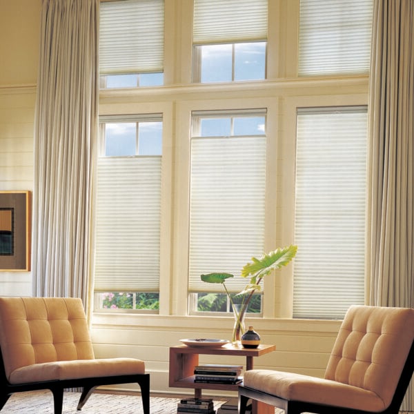A bright living room with large windows, Applause® Honeycomb Shades, two tan cushioned chairs, a wooden side table holding books and a plant, and light-colored curtains framing the windows.