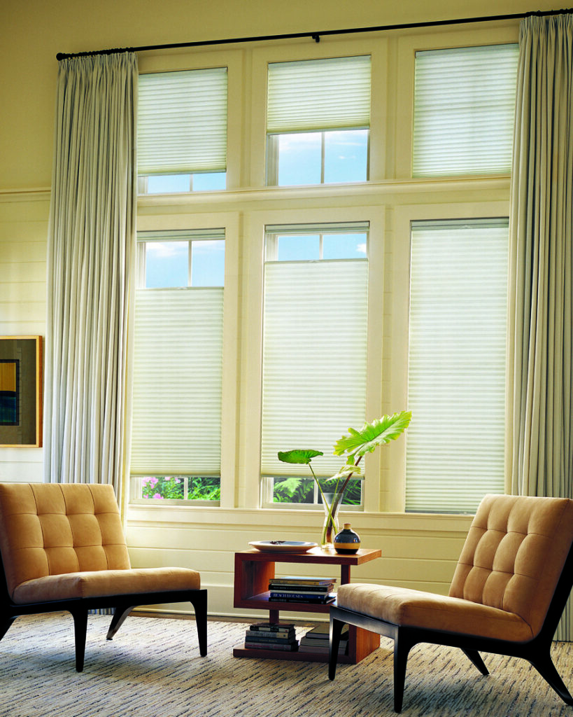 A bright living room with large windows, Applause® Honeycomb Shades, two tan cushioned chairs, a wooden side table holding books and a plant, and light-colored curtains framing the windows.