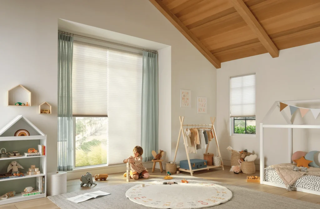 A child plays with toys on a rug in a bright, cozy nursery featuring Duette Honeycomb Shades on the large window, wooden ceiling, small bed with bunting, shelves of toys, and a rack of clothes.