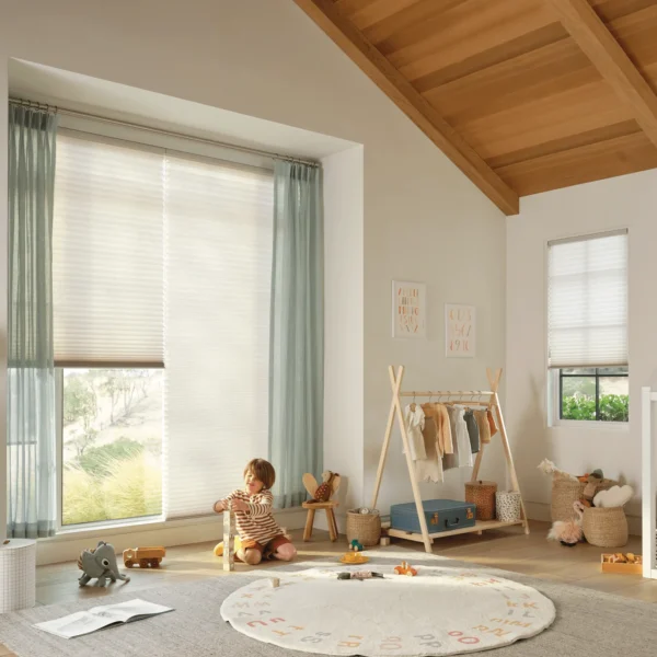A child plays with toys on a rug in a bright, cozy nursery featuring Duette Honeycomb Shades on the large window, wooden ceiling, small bed with bunting, shelves of toys, and a rack of clothes.