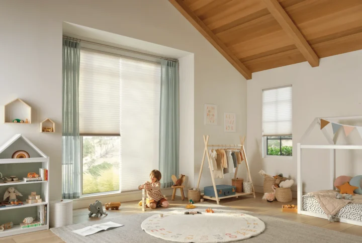 A child plays with toys on a rug in a bright, cozy nursery featuring Duette Honeycomb Shades on the large window, wooden ceiling, small bed with bunting, shelves of toys, and a rack of clothes.
