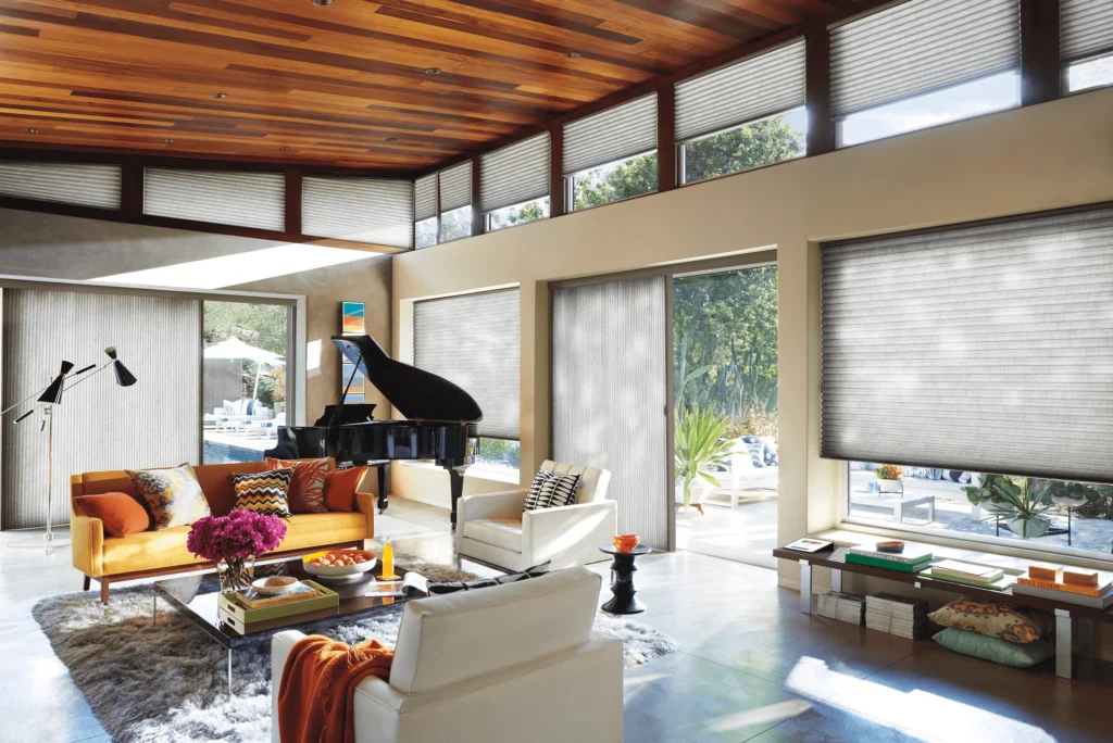 Modern living room with large windows covered by Duette Honeycomb Shades, black grand piano, orange sofa, white armchairs, glass coffee table with flowers, wooden ceiling, sunlight streaming in, and a pool visible outside.