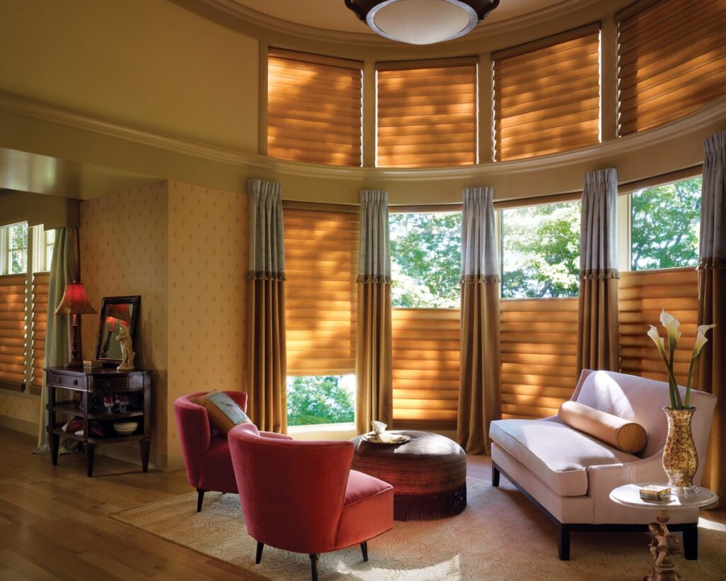 A cozy living room with curved windows features Vignette® Modern Roman Shades, a white sofa, two red chairs, a round coffee table, and decor. Sunlight filters through the shades, creating a warm and inviting atmosphere.