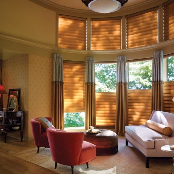 A cozy living room with curved windows features Vignette® Modern Roman Shades, a white sofa, two red chairs, a round coffee table, and decor. Sunlight filters through the shades, creating a warm and inviting atmosphere.