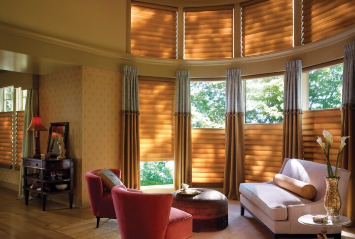 A cozy living room with curved windows features Vignette® Modern Roman Shades, a white sofa, two red chairs, a round coffee table, and decor. Sunlight filters through the shades, creating a warm and inviting atmosphere.