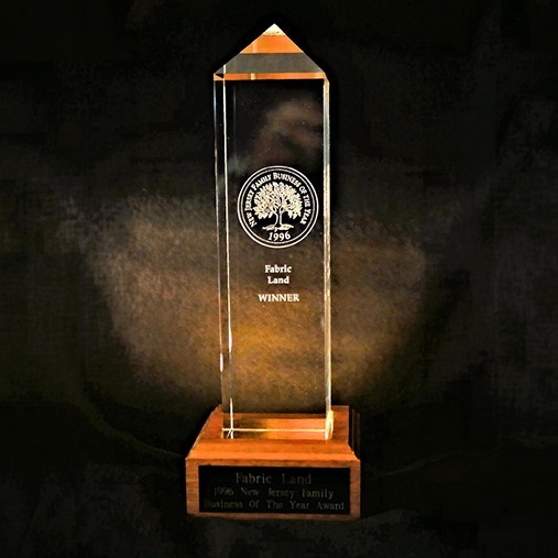 A tall, clear glass obelisk award with a wooden base, engraved with a circular logo and the words "Fabric Land WINNER." The base plaque reads, "1996 New Jersey Family Business of The Year Award.