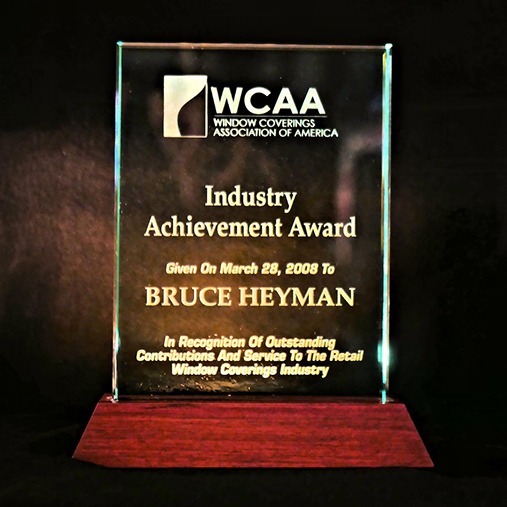 A glass award plaque with a wooden base reads: "WCAA Industry Achievement Award, given on March 28, 2008 to Bruce Heyman, in recognition of outstanding contributions and service to the retail window coverings industry.