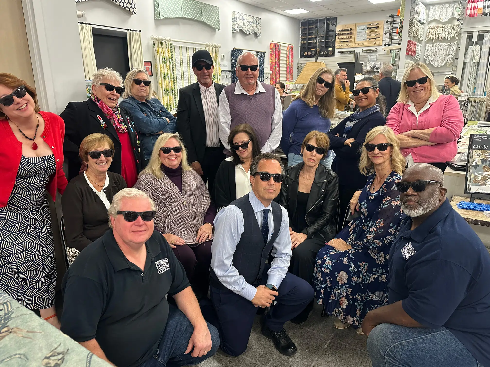 A group of people, both men and women, pose indoors wearing dark sunglasses and smiling. They are in a fabric or craft store, surrounded by bolts of fabric and sewing supplies.