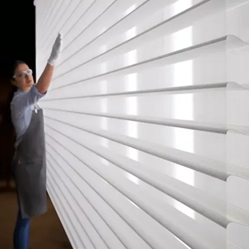 A person wearing safety glasses, gloves, and an apron touches a large, well-lit white louvered surface or panel, possibly inspecting or adjusting it. The scene has an industrial or modern workspace feel.