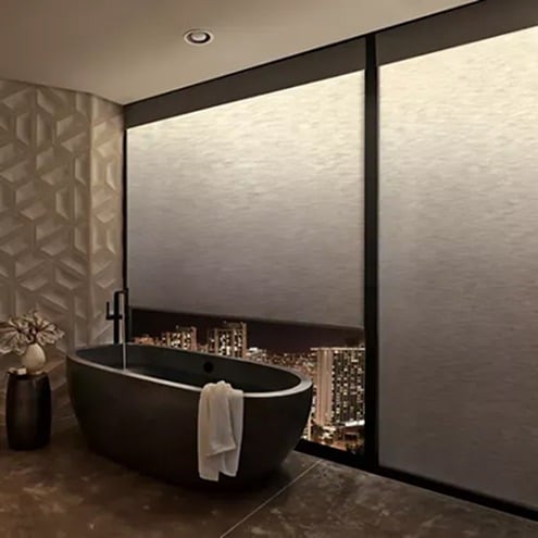 A modern bathroom with a black freestanding bathtub, a white towel draped over the side, geometric wall tiles, and a large window partially covered, revealing a cityscape view at night.