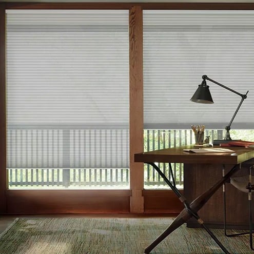 A modern home office features a wooden desk with a black lamp and stationery. Large windows with light-colored blinds let in filtered sunlight, and a green rug covers part of the wooden floor.