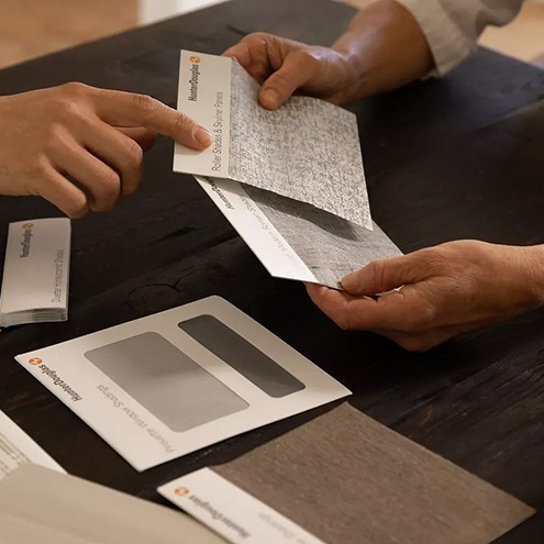 Two people sit at a dark wooden table, examining sample cards with different textures and colors. Several sample envelopes and swatches are spread out on the table.