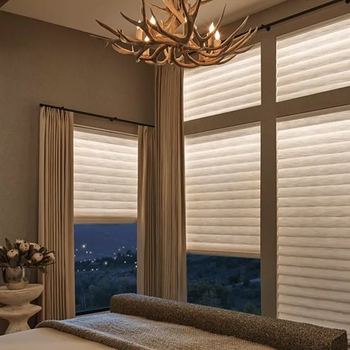 A cozy bedroom with large windows covered by soft, beige shades, letting in diffused light. An antler chandelier hangs from the ceiling above a bed and a side table with a vase of flowers. A nighttime view is visible outside.