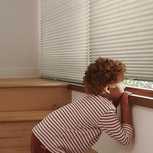 A young child with curly hair and a striped shirt kneels on a wooden bench, peeking through the bottom of closed window blinds with sunlight filtering in.