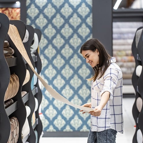 A woman in a plaid shirt smiles as she examines a roll of wallpaper in a store, surrounded by shelves displaying various wallpaper patterns.