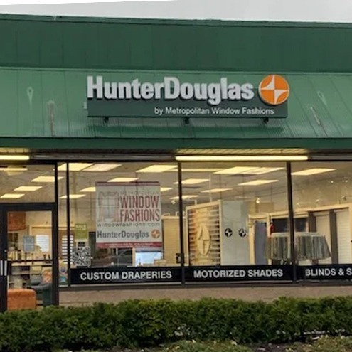 A storefront with a green awning displays the HunterDouglas logo and sign. The windows show blinds, shades, and curtains inside. Text on the glass advertises custom draperies, motorized shades, and blinds & shutters.
