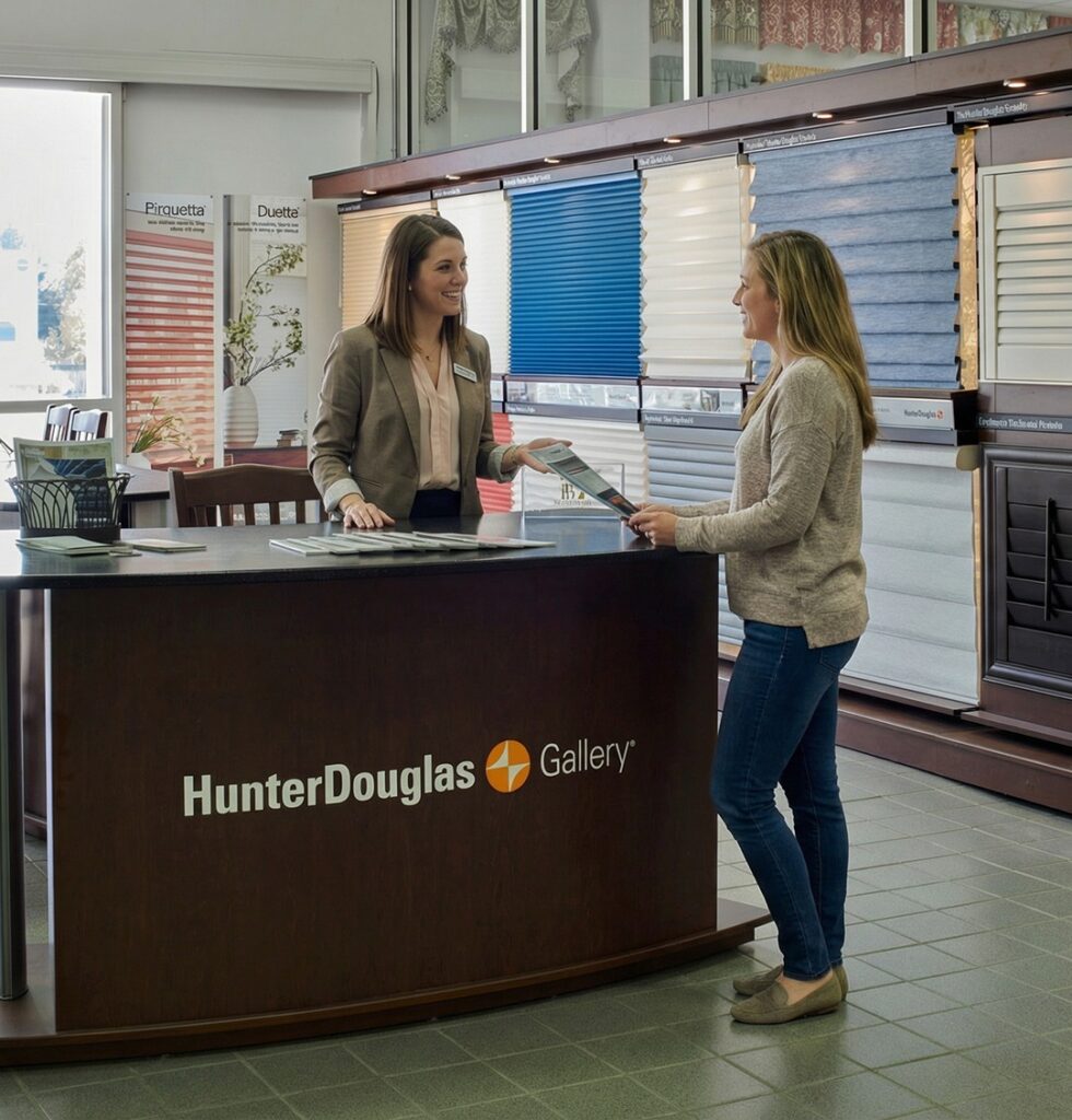 Two women talk at a counter in a Hunter Douglas Gallery showroom, with window blinds and shades displayed behind them. One woman stands and holds brochures while the other stands behind the counter, smiling.
