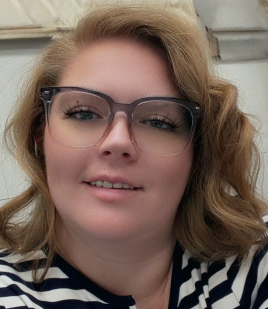 A person with wavy blonde hair, wearing large, clear-framed glasses and a striped shirt, is smiling softly at the camera. The background appears to be indoors with neutral-colored walls.