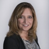 A woman with light brown, shoulder-length hair, wearing a black blazer and patterned top, smiles at the camera against a plain light gray background.