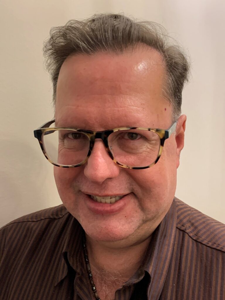 A middle-aged man with short, graying hair is smiling. He is wearing large, tortoiseshell eyeglasses and a brown, striped button-up shirt. The background is plain and light-colored.