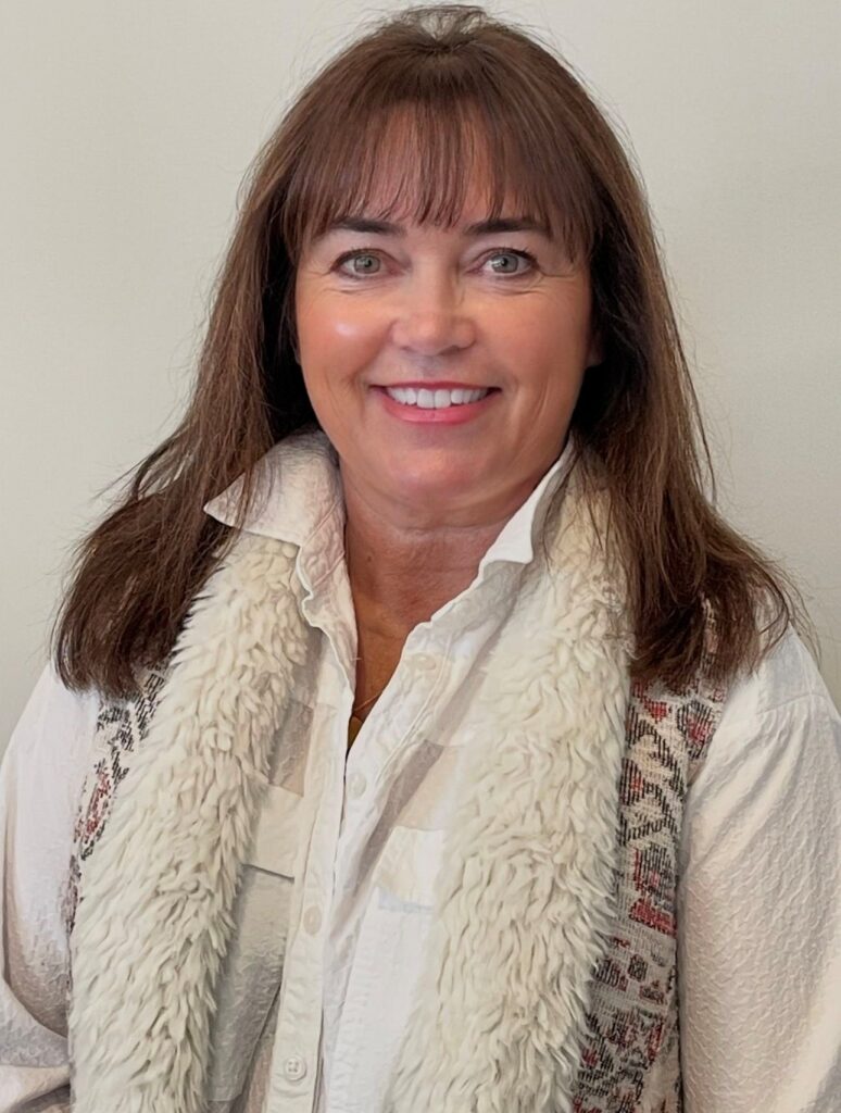 A woman with straight brown hair and bangs is smiling. She is wearing a white shirt and a textured, cream-colored vest with patterned details, standing in front of a plain, light-colored background.