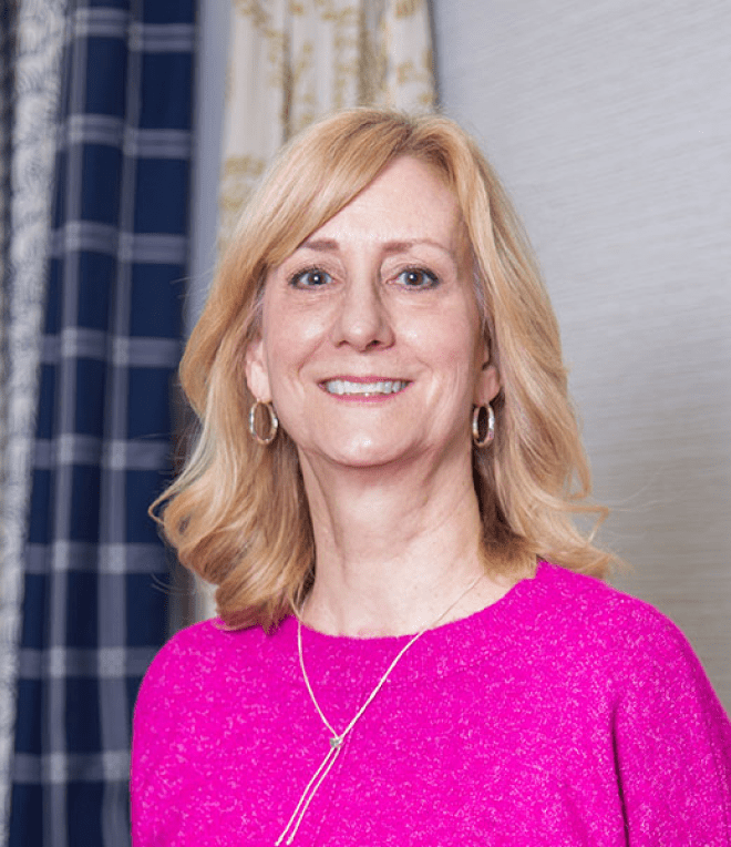 A smiling woman with shoulder-length blonde hair wearing a bright pink top and hoop earrings, standing indoors with patterned curtains in the background.