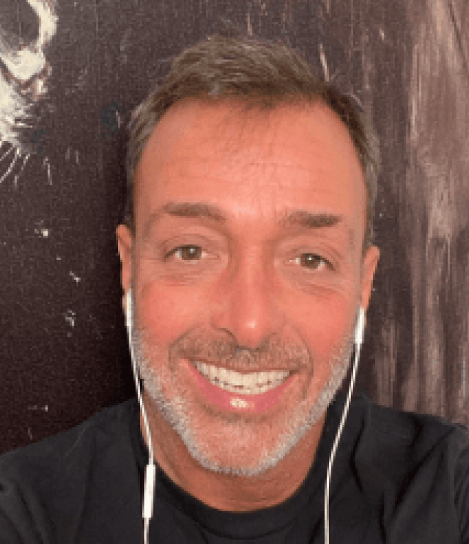 A man with short brown hair and a trimmed beard smiles at the camera, wearing a black shirt and white earphones, with a dark background behind him.