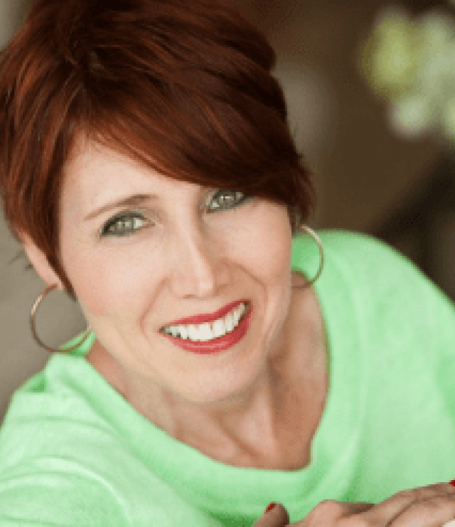 A woman with short auburn hair, wearing large hoop earrings and a light green top, smiles brightly at the camera. The background is softly blurred.