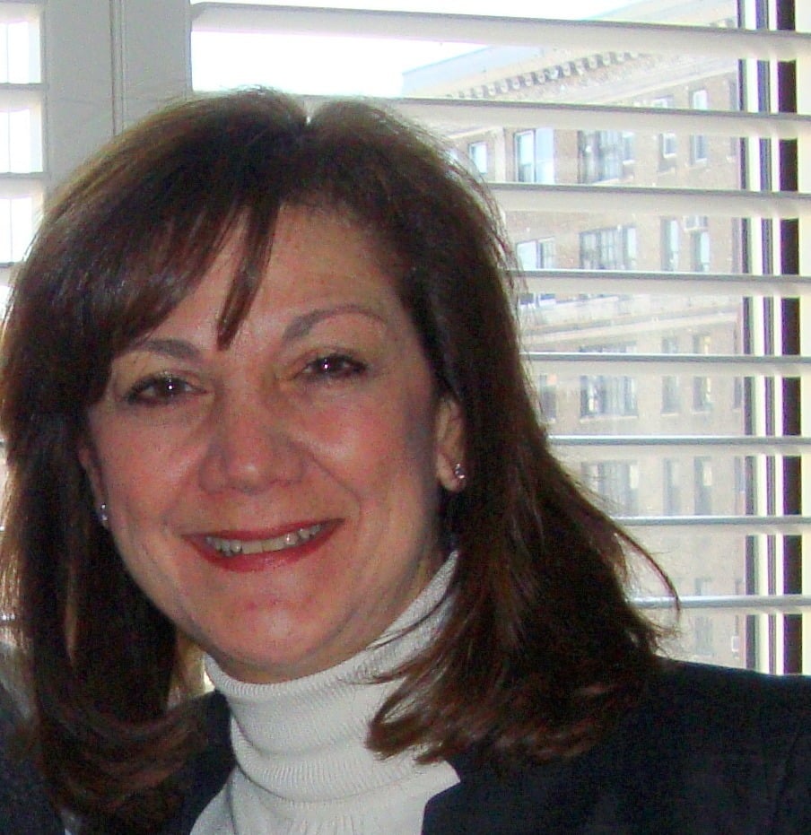 A woman with shoulder-length brown hair and a white turtleneck sweater smiles in front of a window with white blinds and a cityscape visible in the background.