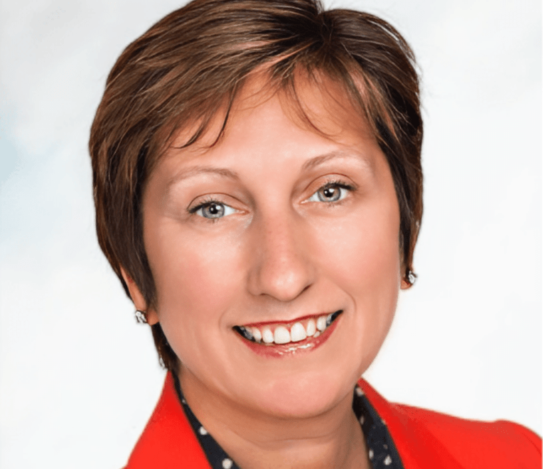 A smiling woman with short brown hair, wearing a red blazer over a dark top with white polka dots, seen against a light, blurred background.