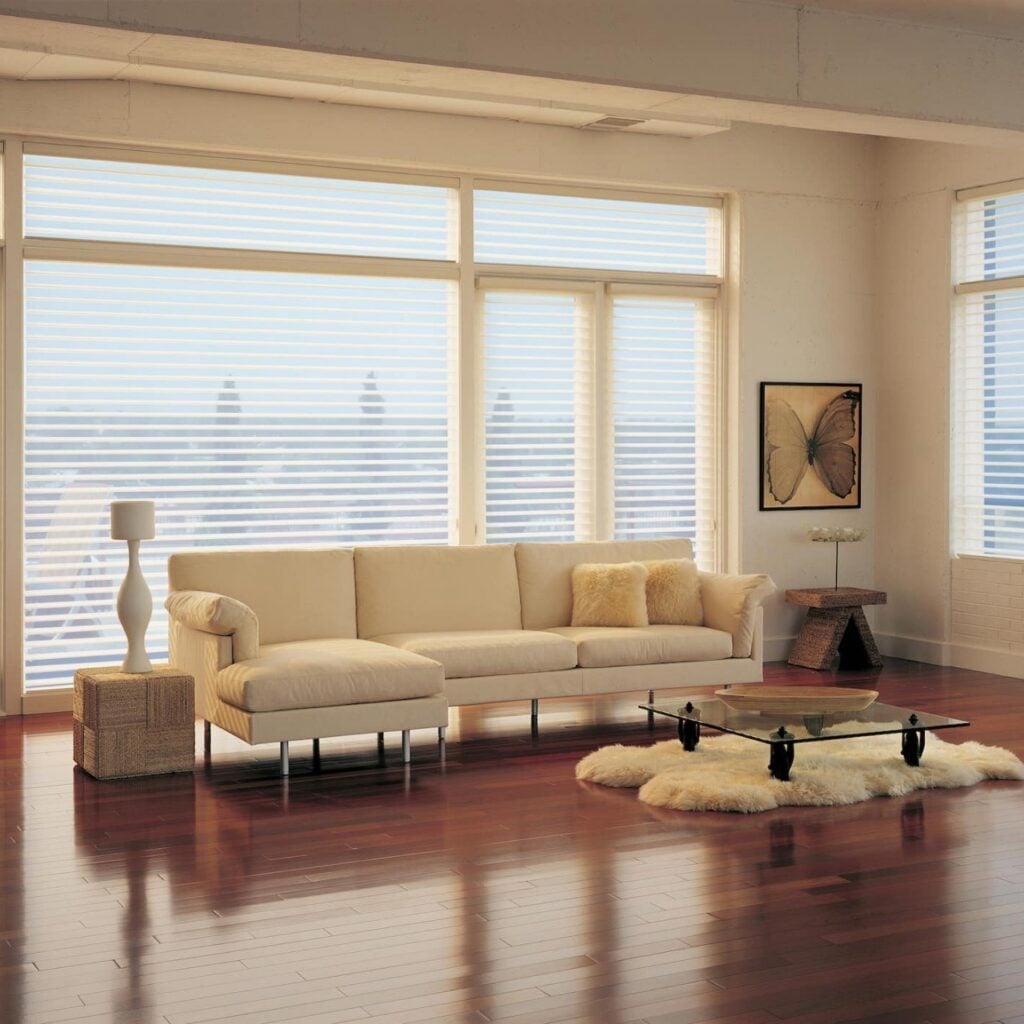 A modern living room with large windows, a cream sectional sofa, fluffy pillows, a glass coffee table on a shag rug, a wicker side table with a lamp, and butterfly artwork on the wall.